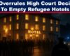 A nighttime protest outside a glowing British hotel, with residents holding placards and police officers and vans present, creating a tense standoff atmosphere.