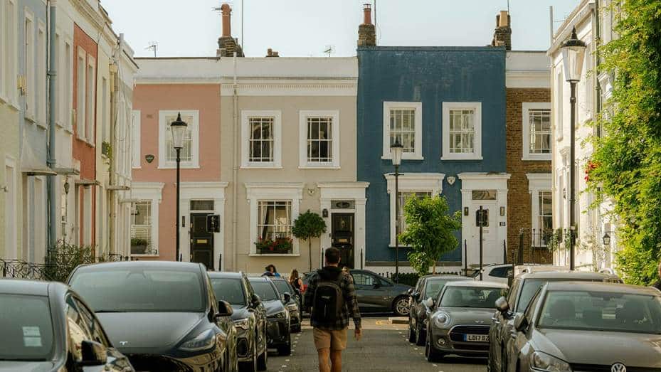 Colorful row of pastel townhouses on a quiet street, with cars parked along both sides and a person with a backpack walking away down the center.