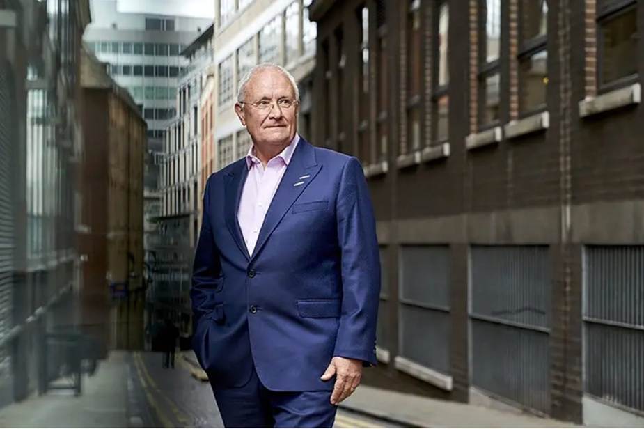 An older man in a navy suit walks down a narrow city alley, hands in pockets, looking ahead.
