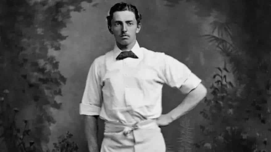 Black-and-white portrait of a man in a white shirt, bow tie, and waist sash posing with hand on hip.