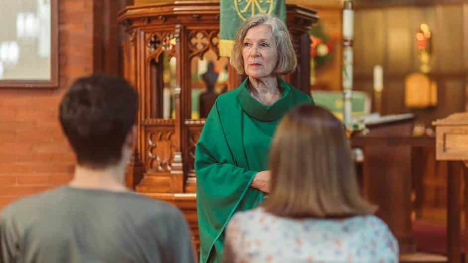 Older woman in a green robe stands at the front of a church, addressing two seated attendees.