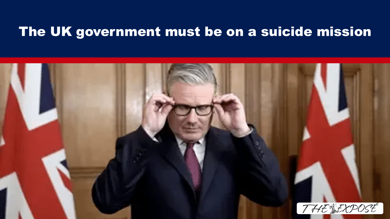 Man in a suit adjusts his glasses in front of British flags, beneath a bold banner that reads: 'The UK government must be on a suicide mission'.