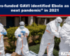 Healthcare workers in full white hazmat suits and blue gloves outdoors, preparing protective equipment near a shrub-lined building.