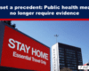City scene with a large red 'STAY HOME' billboard reading 'Essential Travel Only' under a blue header about Covid measures.