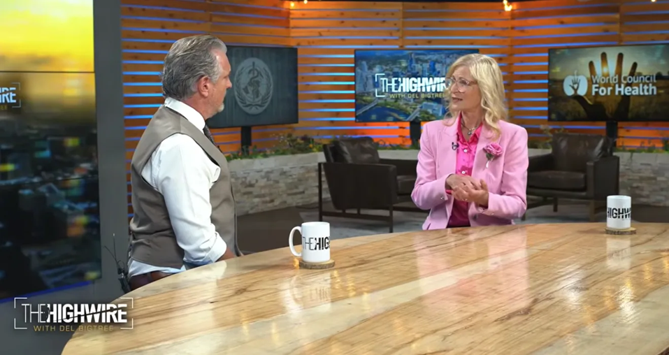 image - The Expose Two people sit at a wooden studio desk during a talk-show interview, a man in a vest facing a woman in a pink blazer across from him.
