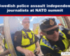 Police officers in high‑visibility vests confront independent journalists at a NATO summit protest, with a crowd in the background and a news logo in the corner