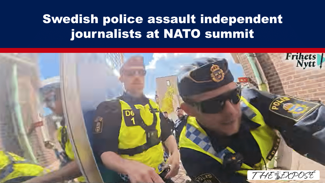 Police officers in high‑visibility vests confront independent journalists at a NATO summit protest, with a crowd in the background and a news logo in the corner