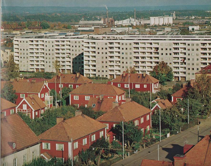 image - The Expose Red wooden houses with red-tiled roofs form a residential foreground, backed by a large white apartment block and distant industrial buildings.