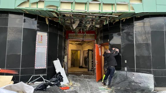 image - The Expose Construction site entrance with exposed ceiling framework, scattered debris, and a worker near an open orange door.
