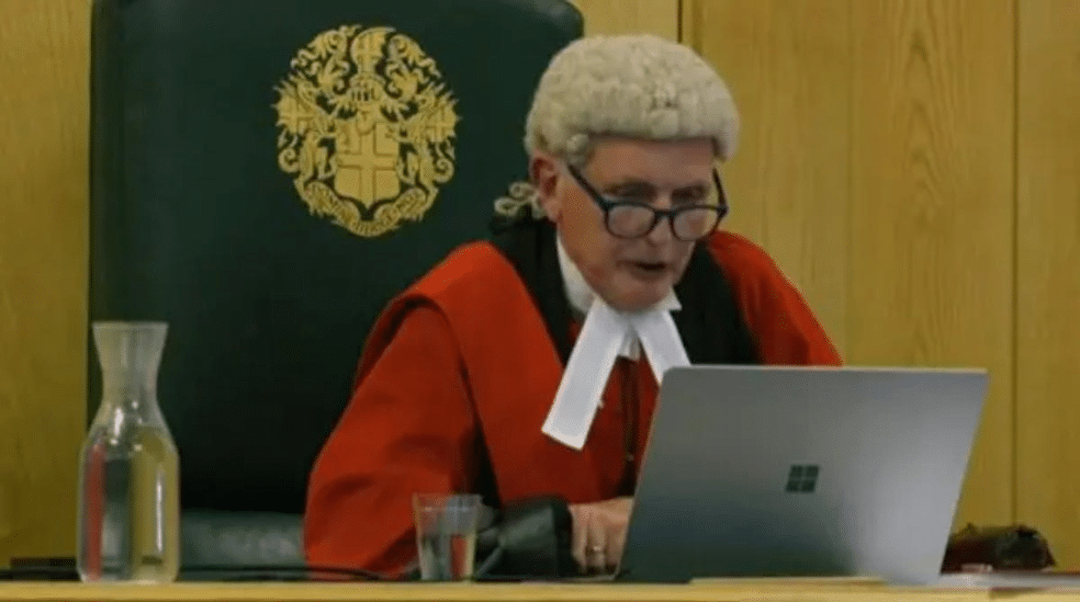 Judge in a red robe and white wig sits at a bench, working on a silver laptop in a courtroom.