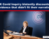 Woman in a suit seated at a curved briefing desk with microphones and papers, a UK Covid-19 Inquiry logo on a blue wall behind her.
