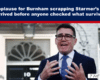 Man in a navy suit and blue tie speaks with animated hand gestures outside a building, beneath a political news banner.