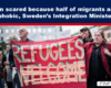 Protesters on a city street hold a large red banner reading 'REFUGEES' as others surround them.