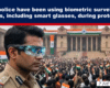 Police officer wearing blue-tinted smart glasses overlooking a large protest with Indian flags and a government building in the background.
