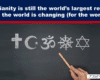 Chalkboard with chalk-drawn symbols of major religions (cross, crescent, Om, wheel, Star of David) and a hand holding chalk beneath a headline about Christianity and global change.